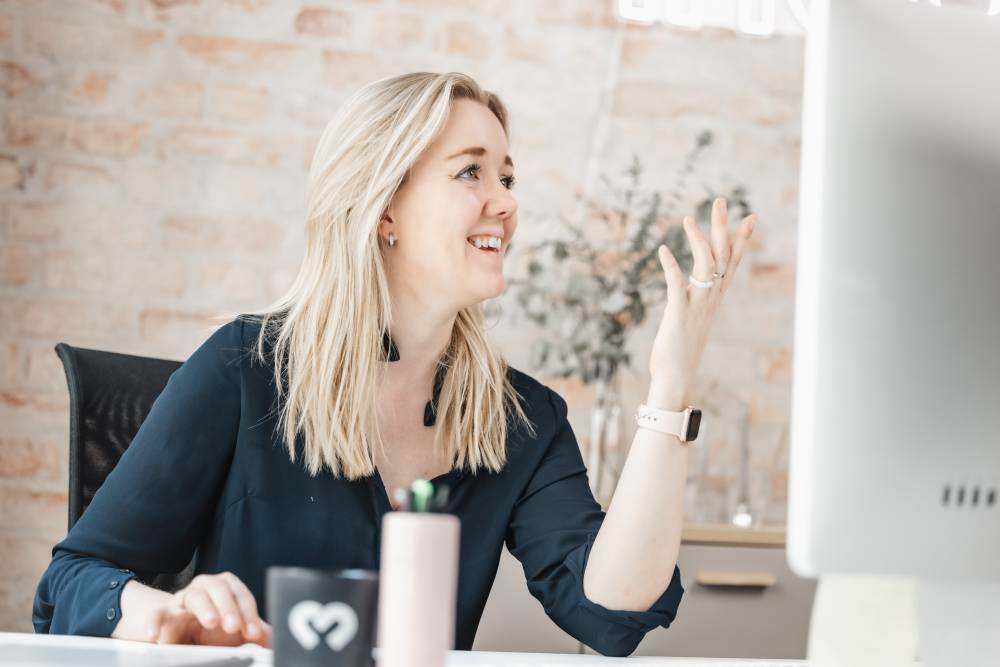 En kvinne med blondt hår sitter ved en dataskjerm og gestikulerer mens hun snakker, i et moderne kontormiljø. Bildet er tatt hos Inbound, et markedsføringsbyrå som spesialiserer seg på B2B-kunder