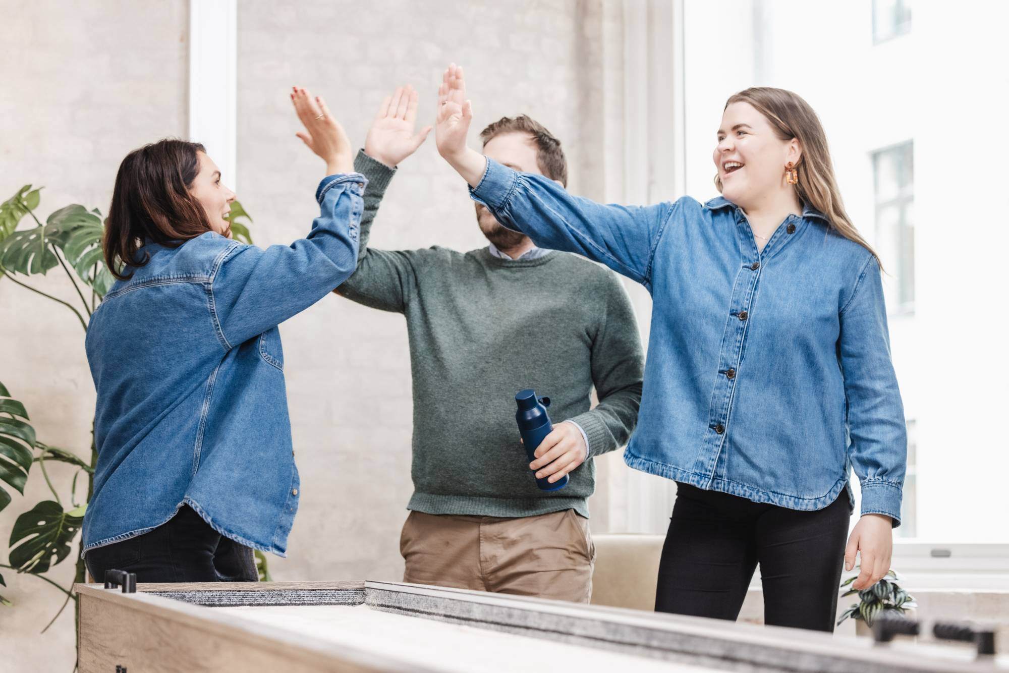 Tre kollegaer feirer et vellykket samarbeid med en high-five i et moderne kontormiljø. Lagånd og suksess i et dynamisk B2B-markedsføringsbyrå.
