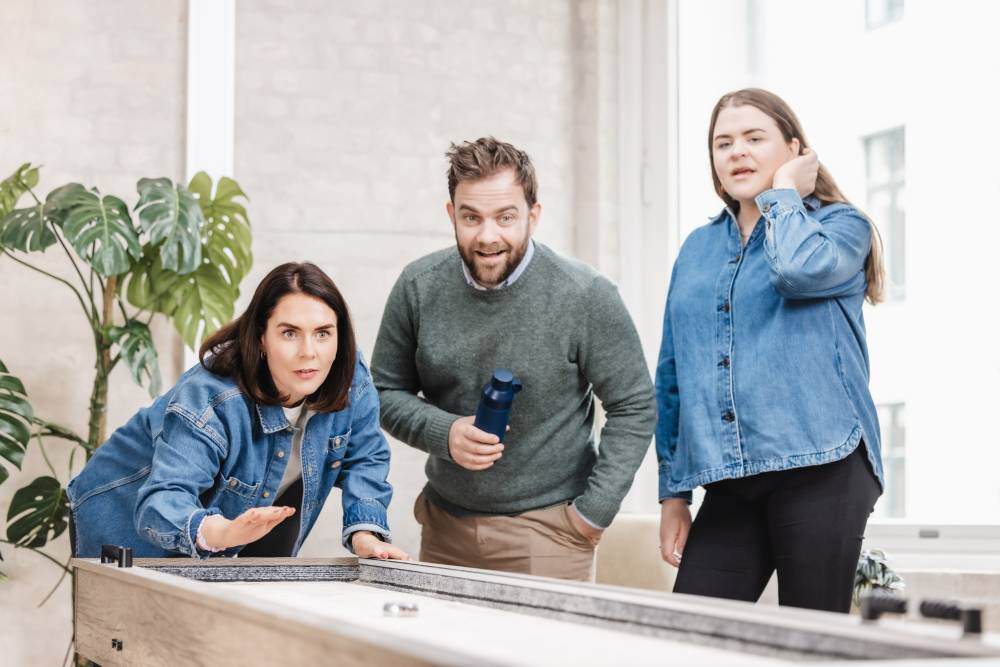 Tre kollegaer fra Inbound, et markedsføringsbyrå som jobber med B2B-kunder, spiller shuffleboard i en avslappet kontorsetting. Stemningen er engasjert og sosial.